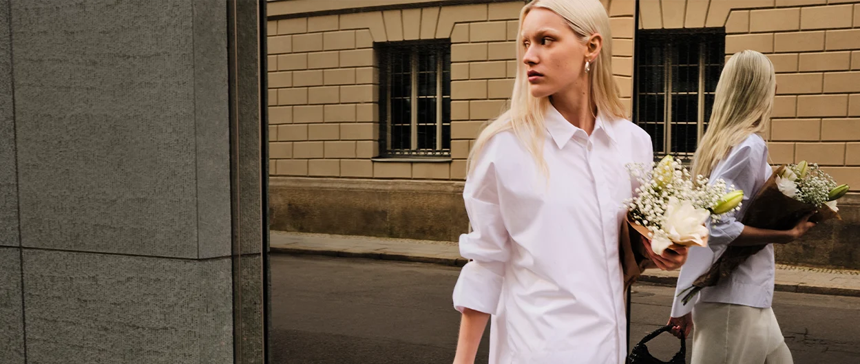 Blonde Frau mit Blumenstrauß geht an einer Spiegelwand entlang, in der ihr Spiegelbild zu sehen ist. (Foto)