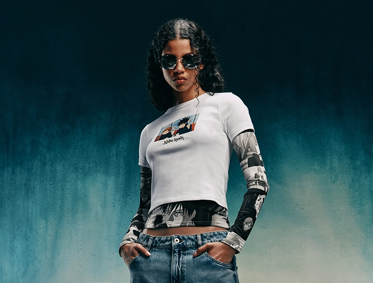 Frau mit Sonnenbrille trägt ein weißes T-Shirt über bedruckten Ärmeln und steht mit Händen in den Hosentaschen vor dunklem Hintergrund. (Foto)